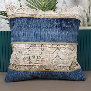 May include: Square decorative pillow with a patchwork design. The pillow features a central section with a faded, patterned beige fabric, bordered by bands of textured blue fabric. The pillow is a home decor item.