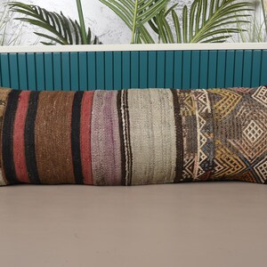 Op de afbeelding: Een lang, rechthoekig kussen met een patchwork-ontwerp. Het kussen heeft een verscheidenheid aan kleuren, waaronder bruin, rood en beige, met geometrische en gestreepte patronen. Het kussen ligt op een neutraal oppervlak.