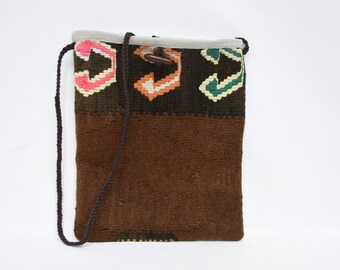 May include: A rectangular shoulder bag with a brown woven body and a patterned top section. The top features geometric designs in pink, orange, and green. The bag has a brown braided strap and a wooden toggle closure. The bag is approximately 10 inches wide and 12 inches tall.
