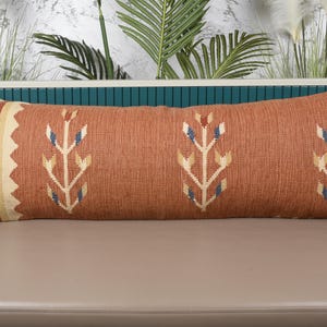 May include: A long, rectangular decorative pillow with a woven design. The pillow features a rust-colored background with cream and blue floral patterns. The left end has a cream and rust striped pattern.