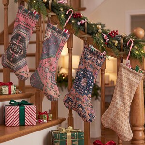 Op de afbeelding: Vier kerstsokken hangend aan een houten trapleuning. Drie sokken hebben ingewikkelde, veelkleurige geometrische patronen in roze, blauwe en grijze tinten, terwijl de vierde is gemaakt van een effen, lichtgekleurde, gestructureerde stof.