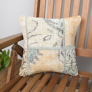 May include: A square throw pillow featuring a patchwork design of woven fabric in muted shades of beige, light orange, and sage green. The textile displays intricate, faded geometric and floral patterns with visible stitching along the seams.