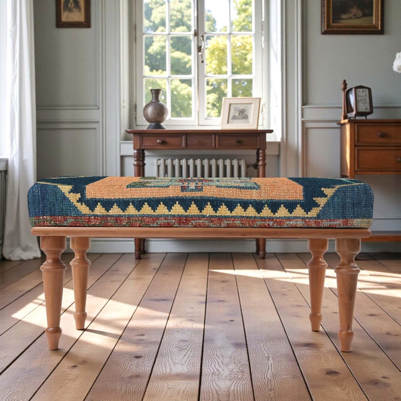 Pu&ograve; includere: Panca in legno con cuscino rettangolare fantasia nei toni del blu, arancione e giallo. La panca ha quattro gambe tornite in legno. Il design del cuscino presenta forme geometriche e un bordo.
