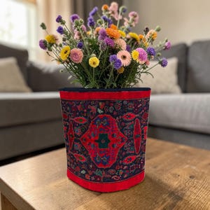 Könnte beinhalten: Ein zylindrischer Blumentopf mit einem gemusterten Stoffdesign, gefüllt mit einem lebendigen Strauß bunter Blumen. Der Topf hat einen roten Rand und einen dunkelblauen oberen Rand. Die Blumen umfassen gelbe, rosa, lila und orangefarbene Blüten.