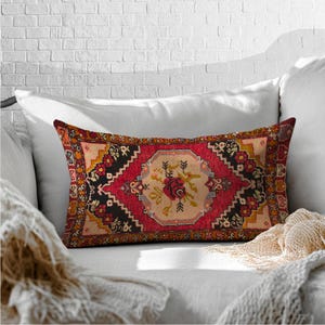 May include: Rectangular decorative pillow with a rug-like design. The pillow features a central floral motif in beige, surrounded by a red border with black and yellow accents. The pillow is placed on a white sofa.