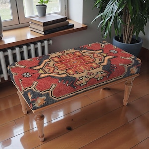 May include: A rectangular bench with a red, black, and tan patterned top, resembling a rug. The bench has light-colored wooden legs. The design on the top is a traditional, geometric pattern. The bench is placed on a wooden floor.