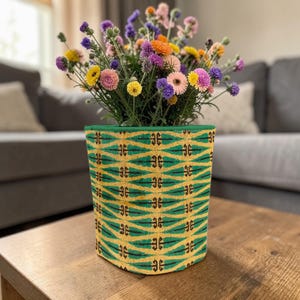 May include: A patterned plant pot with a green rim, featuring a yellow and green geometric design. It contains a colourful arrangement of flowers in shades of pink, yellow, purple, and orange. The pot is placed on a wooden table.