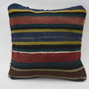 May include: A square throw cushion featuring a woven kilim textile with horizontal stripes in shades of dark blue, mustard yellow, deep red, black, and brown. The fabric has a visible coarse texture with a consistent pattern across the entire surface.