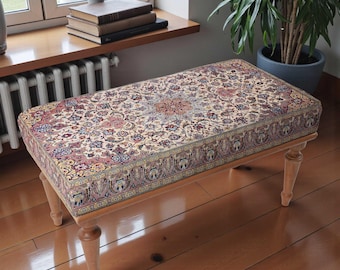 Può includere: Una panca rettangolare con un cuscino a fantasia floreale nei toni crema, rosso e blu. La panca ha quattro gambe in legno di colore chiaro. Il cuscino presenta un design dettagliato e ornato, e la panca è posizionata su un pavimento in legno.