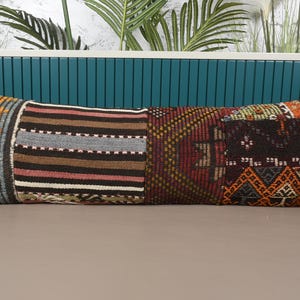 Puede incluir: Un cojín largo y rectangular con un diseño de patchwork. El cojín presenta varios patrones y colores, incluyendo rayas, formas geométricas y una mezcla de tonos rojos, marrones y naranjas. El cojín está diseñado para uso decorativo.