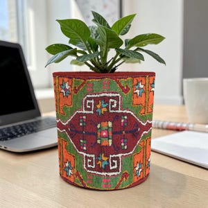Puede incluir: Una maceta cilíndrica con un diseño estampado vibrante. La maceta presenta un borde rojo y un patrón detallado y colorido en tonos rojos, verdes y naranjas. Contiene una planta verde frondosa, creando un acento decorativo.