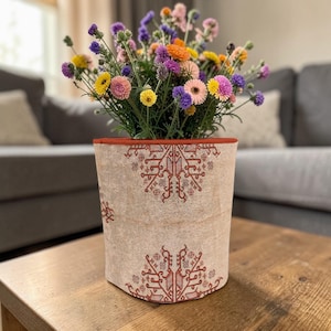May include: A beige cylindrical planter with a red trim and intricate red patterns, filled with a vibrant assortment of colorful flowers. The flowers include yellow, pink, purple, and orange blooms, creating a lively display.