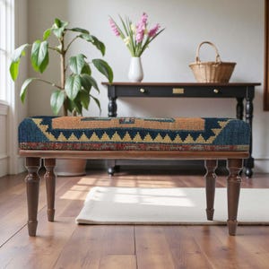 Pu&ograve; includere: Panca in legno con seduta in tessuto fantasia. La seduta presenta un disegno geometrico nei toni del blu, arancione e giallo. La panca ha quattro gambe tornite ed &egrave; posizionata su un pavimento in parquet. Lo stile &egrave; rustico e decorativo. Misura circa 120 cm di lunghezza.