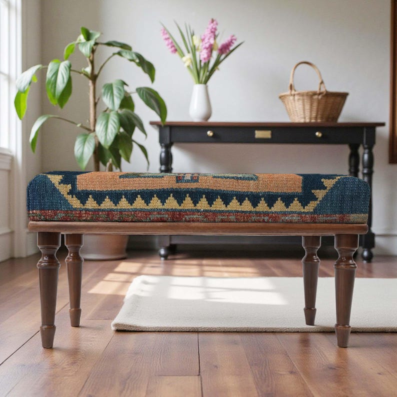 Pu&ograve; includere: Panca in legno con seduta in tessuto fantasia. La seduta presenta un disegno geometrico nei toni del blu, arancione e giallo. La panca ha quattro gambe tornite ed &egrave; posizionata su un pavimento in parquet. Lo stile &egrave; rustico e decorativo. Misura circa 120 cm di lunghezza.