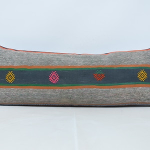 May include: A long, rectangular pillow with a gray background and colorful geometric patterns. The pillow has a dark blue stripe with green and orange stripes on either side. The geometric patterns are in shades of pink, orange, yellow, and green.