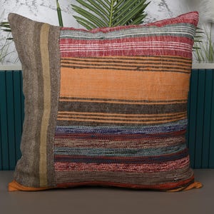 May include: Square decorative pillow with a patchwork design. The pillow features a brown, orange, red, and blue striped pattern. A vertical brown panel is on the left side. The pillow is placed in front of a teal background.