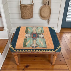 Pu&ograve; includere: Un pouf rettangolare con un top fantasia nei toni dell'arancione, verde acqua e blu navy. Il design presenta forme geometriche e un bordo. Il pouf ha gambe in legno di colore chiaro ed &egrave; posto in una stanza con pavimenti in legno.