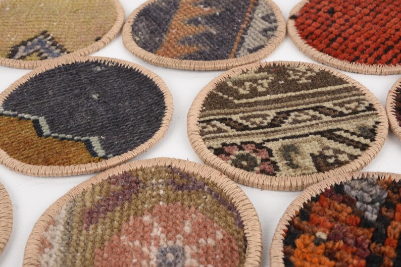 May include: A collection of round coasters with intricate, colorful rug patterns. Each coaster has a beige stitched border. The designs include shades of red, orange, blue, brown, and beige, offering a rustic and decorative touch.