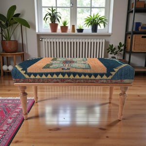Pu&ograve; includere: Un pouf rettangolare con un piano fantasia e gambe in legno chiaro. Il piano presenta un design geometrico nei toni del blu, arancione e giallo. Le gambe sono tornite e hanno una finitura naturale. Il pouf &egrave; posto su un pavimento in legno.