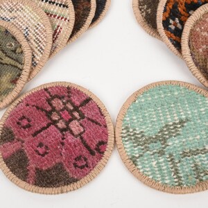 May include: A collection of round coasters with a woven rug design. The coasters feature various colors and patterns, including pink, teal, and brown, with a tan border. The coasters are arranged on a white surface.