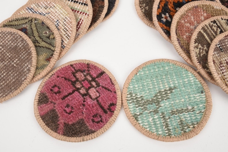 May include: A collection of round coasters with a woven rug design. The coasters feature various colors and patterns, including pink, teal, and brown, with a tan border. The coasters are arranged on a white surface.