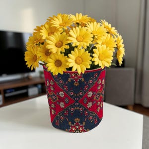 Op de afbeelding: Een bloemstuk van gele madeliefjes in een decoratieve vaas. De vaas heeft een rood en marineblauw patroon. De bloemen staan in volle bloei en creëren een vrolijke uitstraling.