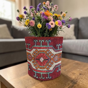 May include: A burgundy fabric planter with a detailed geometric pattern in orange, teal, and white holds a vibrant bouquet of colourful flowers. The flowers include yellow, pink, purple, and orange blooms. The planter sits on a wooden table.