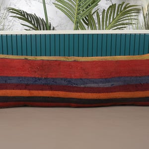 May include: A long, rectangular pillow with a woven, striped design. The pillow features bands of red, blue, orange, and black. The pillow's ends have a faded appearance, and the overall aesthetic is rustic and textured.