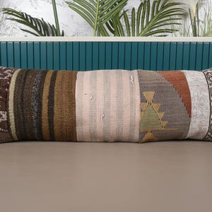 May include: A long, rectangular pillow with a patchwork design. The pillow features various textured fabrics in shades of brown, beige, and gray, with a section of pink and a geometric pattern. The pillow is approximately 40 inches long.