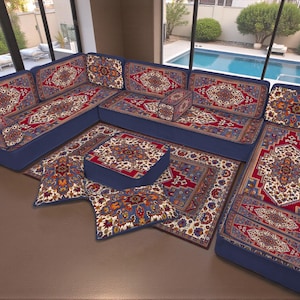 Può includere: Un divano componibile con struttura blu e cuscini con un motivo tradizionale a tappeto in rosso, crema e oro. Il set include un divano angolare, un pouf quadrato e cuscini decorativi, creando un'area salotto accogliente.