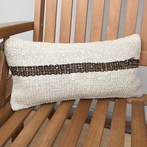 May include: A white woven throw pillow with a brown woven stripe across the center. The pillow is sitting on a wooden chair.