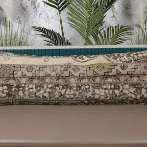 Puede incluir: Cojín decorativo rectangular con un diseño vintage. El cojín presenta un fondo beige con patrones geométricos y florales marrones, rosas y verdes. El cojín está colocado sobre una superficie neutra con vegetación en el fondo.