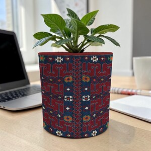 Op de afbeelding: Een cilindrische plantenpot met een gedetailleerd patroon in rood, blauw en oranje. De pot bevat een groene plant met bladeren. De pot staat op een houten bureau naast een laptop.
