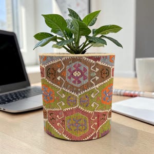 May include: A cylindrical plant pot with a colorful, patterned design. The pot features a geometric pattern in shades of brown, green, orange, and purple. The pot holds a green leafy plant. The pot is sitting on a wooden desk.