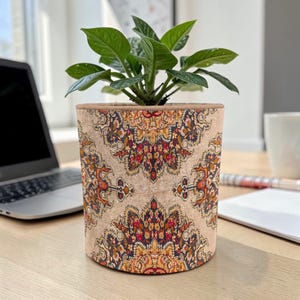 May include: A beige plant pot with a detailed, colorful pattern, holding a small green plant. The pot's design features red, orange, and brown floral motifs. The pot is sitting on a wooden desk, next to a laptop.