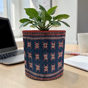 May include: A potted plant in a decorative fabric pot. The pot is navy blue with a repeating pattern of red and white floral designs. The rim of the pot is red. The plant has green leaves.