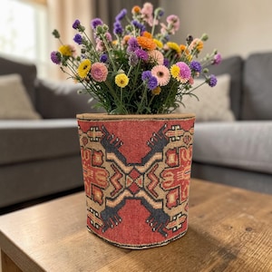 Può includere: Una vivace composizione floreale in una fioriera decorativa con motivi rossi e beige. La fioriera è su un tavolo di legno. I fiori includono fiori gialli, rosa, viola e arancioni. Lo sfondo include un divano grigio.