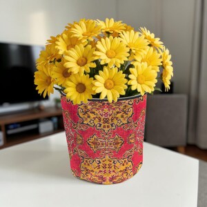 May include: A vase of yellow daisies, set in a red and gold patterned container. The vase is cylindrical and sits on a white table. The flowers and vase create a striking contrast. The vase's pattern is reminiscent of a traditional rug.