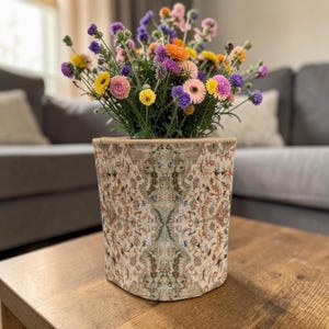 May include: A decorative flower pot with a beige and multi-colored oriental rug design. The pot is filled with a vibrant arrangement of colorful flowers, including yellow, pink, purple, and orange blooms. The pot sits on a wooden table.