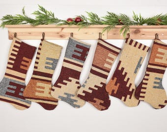 Könnte beinhalten: Sechs Weihnachtsstrümpfe mit geometrischen Mustern in Beige, Braun und Grau hängen an einem Holzregal. Das Regal ist mit Grün und roten Ornamenten dekoriert und schafft eine festliche Atmosphäre.