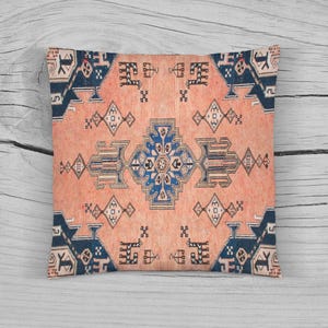 Puede incluir: Cojín cuadrado con fondo melocotón y un patrón geométrico en azul marino y crema. El cojín presenta un diseño de medallón central y motivos repetidos en los bordes, que se asemejan a una alfombra tradicional.