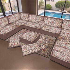 Puede incluir: Un sofá seccional con un estampado floral en tonos beige, rojo y marrón. El sofá está dispuesto en forma de L, con cojines a juego y una otomana rectangular. Una alfombra estampada se encuentra debajo de los muebles.