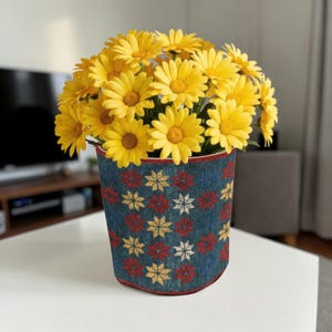 Op de afbeelding: Een boeket felgele madeliefjes in een decoratieve bloempot. De pot heeft een patroon van rode, gele en witte bloemen op een denim-stijl achtergrond. De pot heeft een rode rand aan de boven- en onderkant.