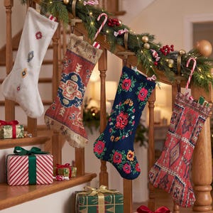 Op de afbeelding: Vier kerstsokken hangend aan een houten trapleuning. Eén is wit met klein geometrisch borduurwerk, één heeft een rood en blauw sierlijk tapijtpatroon, één is donkerblauw met bloemmotieven en één heeft een rood geometrisch patroon.