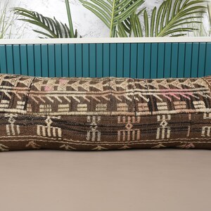 Puede incluir: Un cojín rectangular decorativo con un patrón geométrico marrón y beige. El cojín tiene una forma larga y estrecha y está hecho de una tela tejida. El diseño incluye formas y líneas repetidas en tonos marrones, beige y rosas.