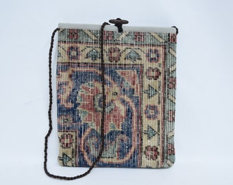 May include: A rectangular shoulder bag with a woven rug design. The bag features a repeating pattern in shades of blue, red, and beige. It has a brown rope strap and a wooden toggle closure at the top.