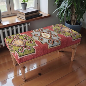 May include: A rectangular upholstered bench with a red, green, and gray patterned top. The design features geometric shapes and floral accents. The bench has four light-colored wooden legs.