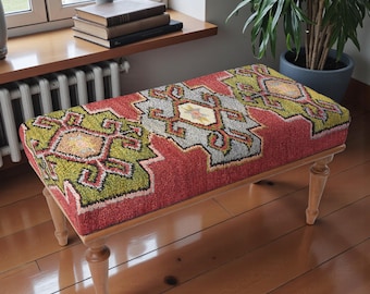 Può includere: Una panca rettangolare imbottita con un piano a fantasia rosso, verde e grigio. Il design presenta forme geometriche e accenti floreali. La panca ha quattro gambe in legno di colore chiaro.