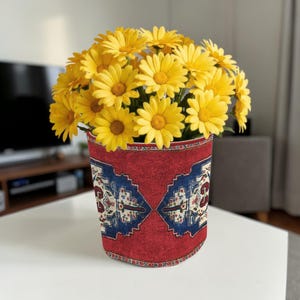 May include: A vase of yellow daisies, presented in a red vase with a blue and white patterned design. The vase is cylindrical and rests on a white surface, offering a striking contrast.