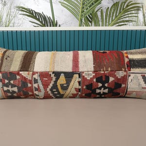 May include: Rectangular decorative pillow with a patchwork design. The pillow features various geometric patterns in shades of red, brown, beige, and black. The pillow is placed on a neutral surface with a teal background.